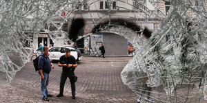Zwei Männer sind im Hamburger Schanzenviertel auf der Straße durch eine zerbrochene Glasscheibe hindurch zu sehen