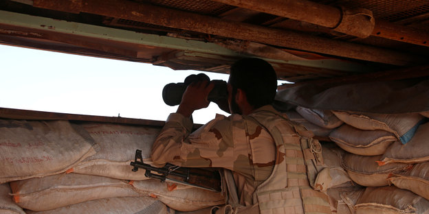 Uniformierter und bewaffneter Mann schaut durch ein Fernglas aus einem Gefechtsstand