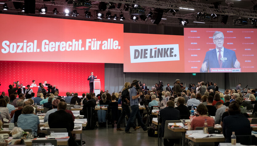 Viele Leute in einer Halle, im Hintergrund ein Schild: "soial. gerecht. für alle. die linke"