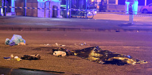 eine Leiche unter einem schwarzen Plastisack auf einer Straße, daneben weitere Gegenstände