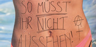 Auf einem Werbeplakat, das eine Frau im Bikini zeigt, steht per Hand geschrieben: "SO MÜSST IHR NICHT AUSSEHEN!"