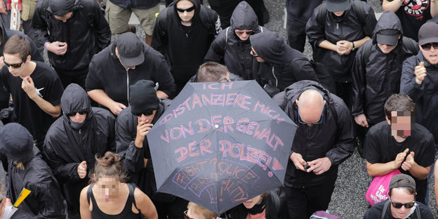 Schwarz gekleidete DemonstrantInnen tragen einen Regenschirm, auf dem steht: „Ich distanziere mich von der Gewalt der Polizei“