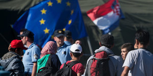 Menschen mit Rucksäcken stehen vor Polizisten und Fahnen von Europa und Kroatien