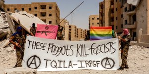 zwei Menschen in Tarnanzügen zeigen ein Transparent mit der Aufschrift "These faggits kill fascists", darüber eine Regenbogenfahne, dahinter eine halb zerstörte Stadt