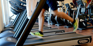 Fitnessstudio von innen. Viele Laufbänder nebeneinander, auf dem vordersten sieht man die Beinen eines weißen Sportlers.