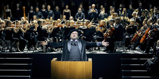ein Mann steht mit ausgebreiteten Armen am Rednerpult, im Hintergrund ein Orchester