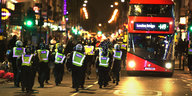 Polizisten in voller Montur laufen auf einer nachts erleucheten Straße, im Hintergrund ein großer roter Bus