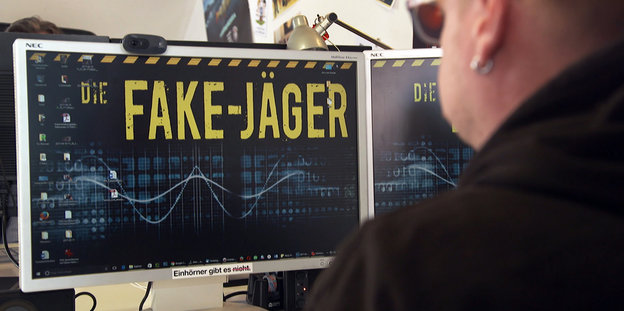 Ein Mann sitzt vor einem großen Computer-Bildschirm, auf dem in gelber Schrift "Fake-Jäger" steht