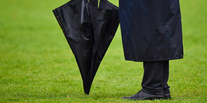 Ein Mann in Schwarz steht auf einem perfekten Rasen, vor ihm ein schwarzer Regenschirm