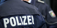 Der Rücken eines Polizisten, darauf der Schriftzug "POLIZEI". Im Hintergrund ein weiterer Polizist.