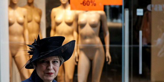 Therasa May steht vor einem Schaufenster mit nackten Schaufensterpuppen, darüber ein Schild auf dem Alles 60 Prozent steht