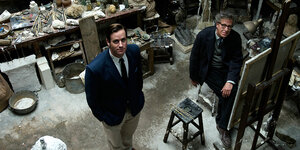 Zwei Männer stehen in einem Atelier, schauen nach oben, in die Kamera, hinter ihnen viele Skulpturen.