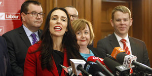 Eine Frau in roter Bekleidung steht vorne am Pult mit Mikros - hinter ihr Männer und eine weitere Frau