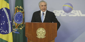 Ein älterer Mann in Anzug steht an einem Rednerpult, neben ihm die brasilianische Flagge