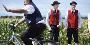 Zwei Männer in Tracht stehen vor einer Wiese, davor fährt ein Mann auf dem Fahrrad vorbei