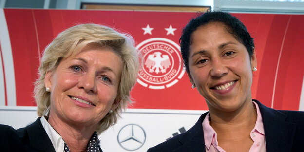 zwei lächelnde Frauen vor einem großen DFB-Logo