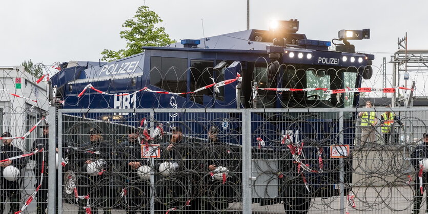 ein Wasserwerfer hinter einem mit Natodrahrt bewehrten Zaun