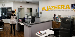 Eine Frau geht durch ein Büro mit abgetrennten Sitzplätzen und dem Schriftzug "Al Jazeera" im Hintergrund