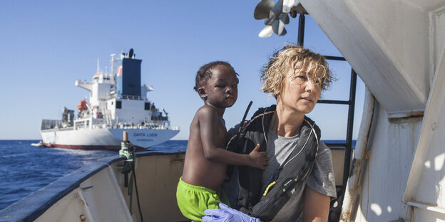Eine blonde Frau an Bord eines Schiffes hält ein schwarzes Baby auf dem Arm