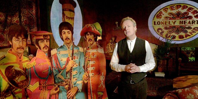 Figuren der Beatles im Hintergrund. Davor der Musiklehrer