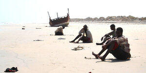 Mehrere Männer sitzen unter der gleißenden Sonne am Strand, in der Ferne ein Schiffswrack