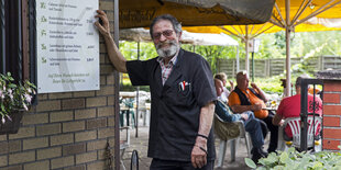 Nicolaos Bitas stützt sich mit der Hand auf das Menü, das im Biergarten seiner Kneipe an der Wand hängt