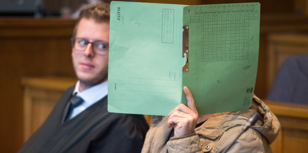 Ein Mann in einem Gerichtssaal hält eine Mappe vor sein Gesicht
