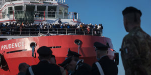 Soldaten vor einem roten Schiff auf dem viele Flüchtlinge stehen