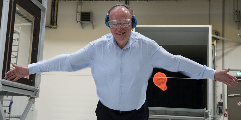 Ein Mann in weißem Hemd hat eine Schutzbrille und Ohrenschützer auf und streckt beide Arme zur Seite