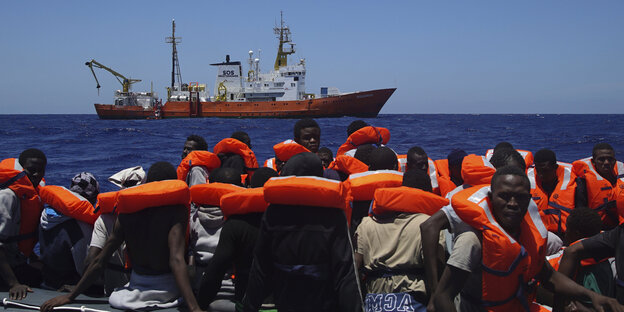 Menschen mit Rettungswesten auf einem Schlauchboot im Meer, in einiger Entfernung fährt ein Schiff