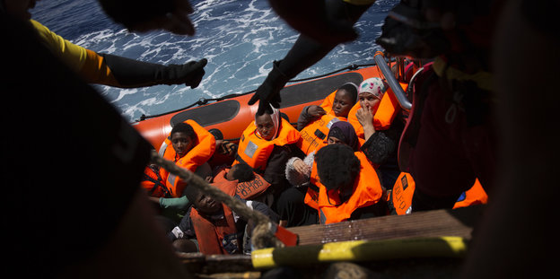 Menschen in einem Schlauchboot, davor als Silhouetten HelferInnen