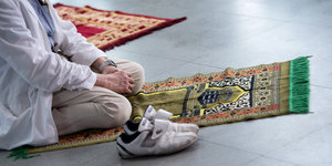 Eine Person kniet auf einem Gebetsteppich und hat ihre Turnschuhe neben ihm abgestellt.