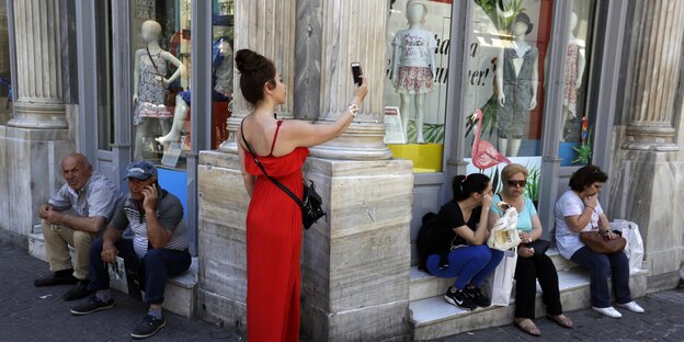 Passanten sitzen vor einem Laden, eine Frau im roten Abendkleid steht vor ihnen und macht ein Selfie von sich