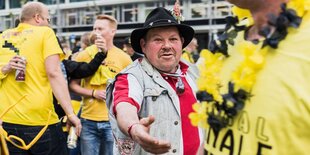 Männer in Fußballtrikots stehen auf einem Platz. In der Mitte steht ein Mann mit Hut, der einem anderen seine Hand entgegenstreckt.