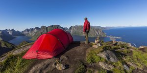 Jemand steht neben seinem Zelt auf einer Bergspitze. Am Horizont sind Wasser und andere Felsformationen zu sehen.