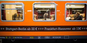 In einem orangenfarbenen Zugabteil sieht man zwei Reisende, auf dem Zugabteil steht "Stuttgart-Berlin 22 Euro / Frankfurt-Hannover 13 Euro"
