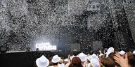 Menschen stehen mit weißen Hüten vor einer Bühne, über ihnen schweben weiße Teilchen in der Luft