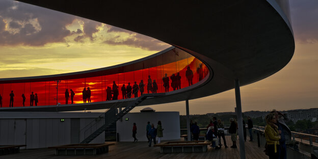 Eine geschwungene, verglaste Brücke mit rot getönten Scheiben vor Sonnenuntergang mit Menschen
