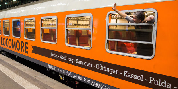 Ein Mann winkt aus dem Fenster eines orangefarbenen Zuges