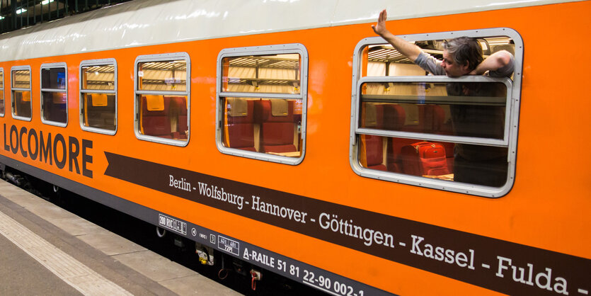 Ein Mann winkt aus dem Fenster eines orangefarbenen Zuges