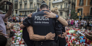 Ein Polizist ist von hinten zu sehen, er umarmt zwei Menschen, im Hintergrund sind Kerzen und Blumen aufgestellt