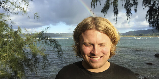 ein grinsender Mann am Ufer, hinter ihm ein Regenbogen