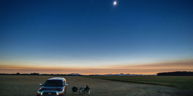 Auf einem großen Feld steht ein Auto und zwei Menschen sitzen auf Stühlen und blicken in die totale Sonnenfinsternis am Himmel