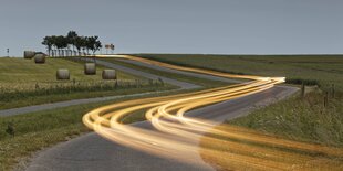 Bei Dämmerung läuft eine Lichtspur über eine Landstraße.