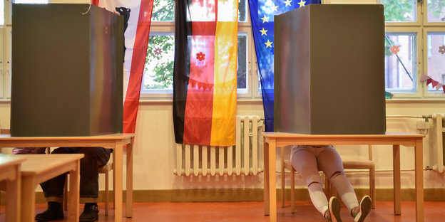 Zwei Wahlkabinen, aus denen Beine hervorschauen, dazwischen die Fahnen von Berlin, Deutschland, Europa