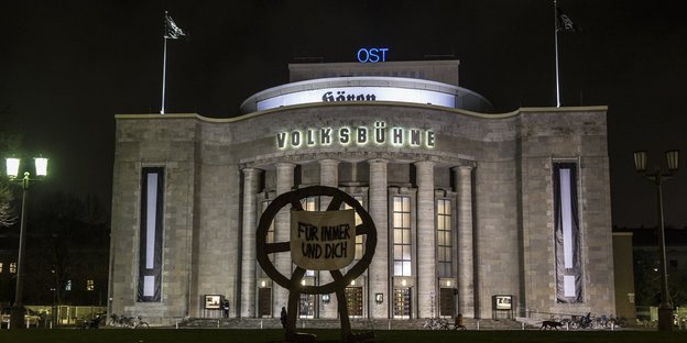 Die Berliner Volksbühne bei Nacht und beleuchtet. An der Radskulptur vor dem Haus ein Transpi mit der Aufschrift "Für immer und Dich"