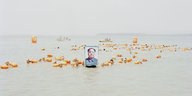 Ein Fluss, in dem Menschen mit orangefarbenen Schwimmhilfen schwimmen. Sie halten ein Porträt von Mao Zedong in die Höhe