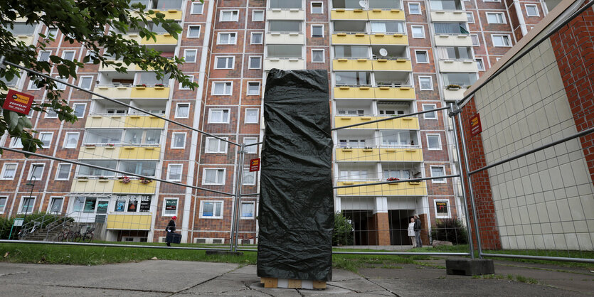 Vor einem Plattenbau steht eine verhüllte Säule. Sie ist mit einem Gitter abgesperrt