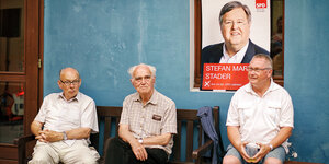 drei alte Männer sitzen auf einer Bank vor einer Wand, darauf ein SPD-Plakat mit dem Gesicht von Klaus Maria Stader