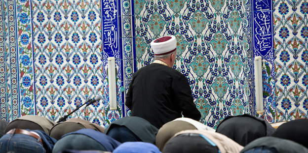 In einer Moschee haben sich Menschen zum Gebet auf Knien verbeugt, vor blau gekachelten Wänden kniet ein Geistlicher